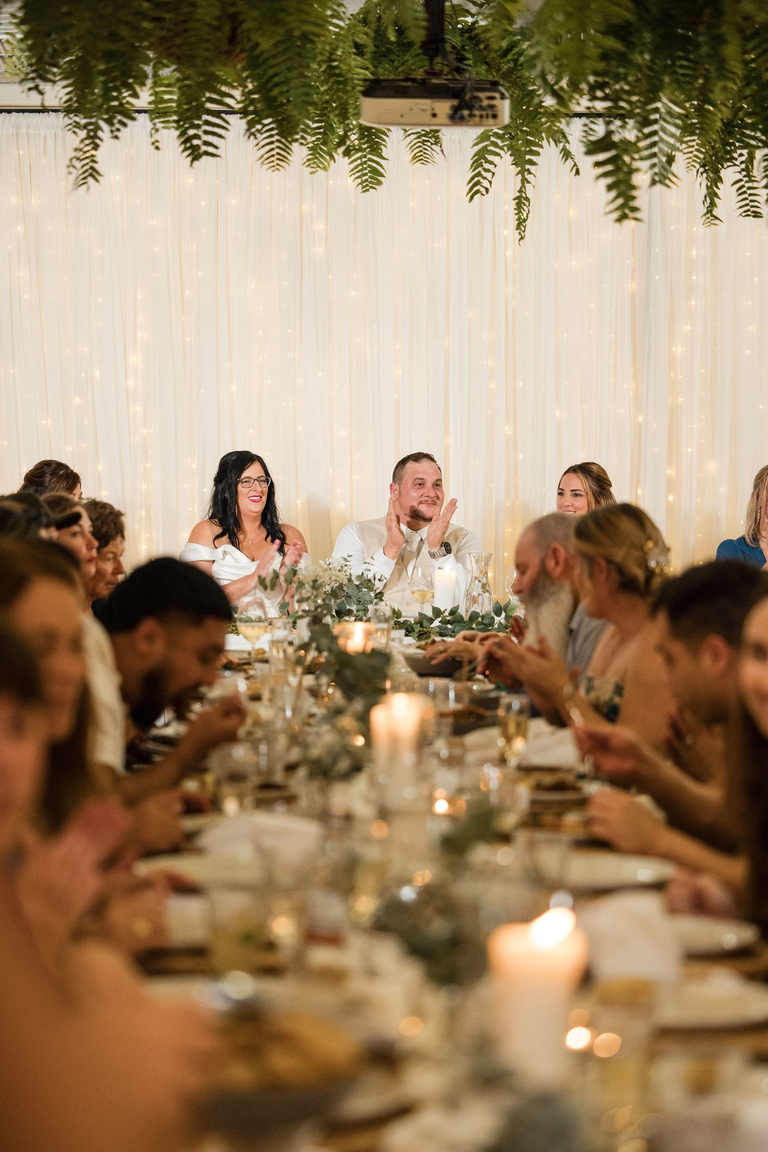 Elegant wedding reception with bride and groom seated at candlelit table, fairy lights and fern garlands decorating the venue.