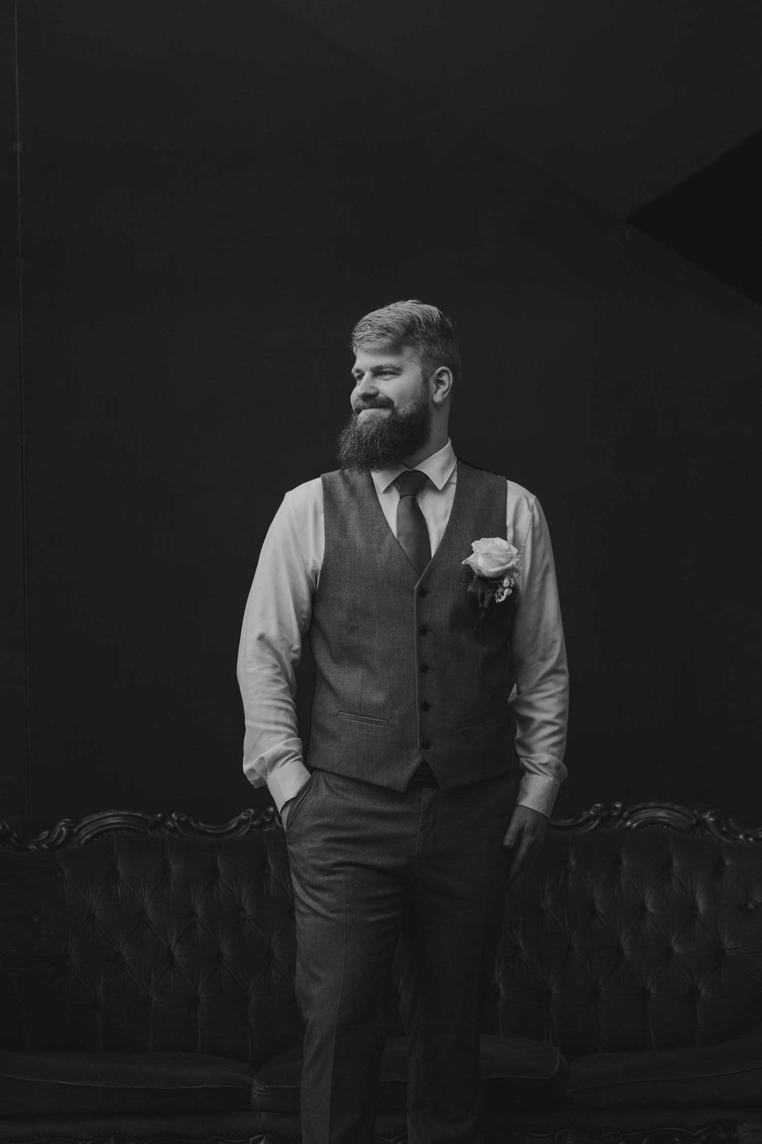 Bearded groom in vest and tie with boutonniere poses confidently in black and white portrait photo.