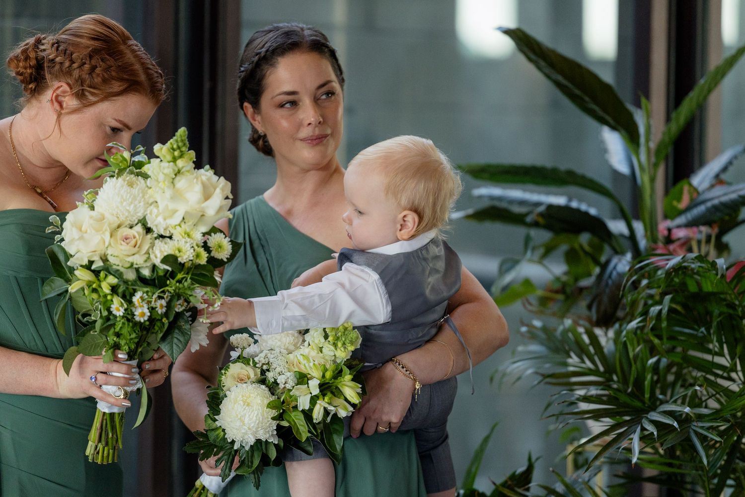 Two bridesmaids in sage green dresses hold white bouquets while one carries a baby in a grey suit at a wedding.