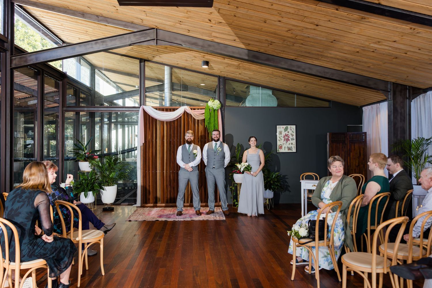 Wedding ceremony in a modern indoor venue with wood ceiling, guests seated in bentwood chairs facing couple at floral backdrop.