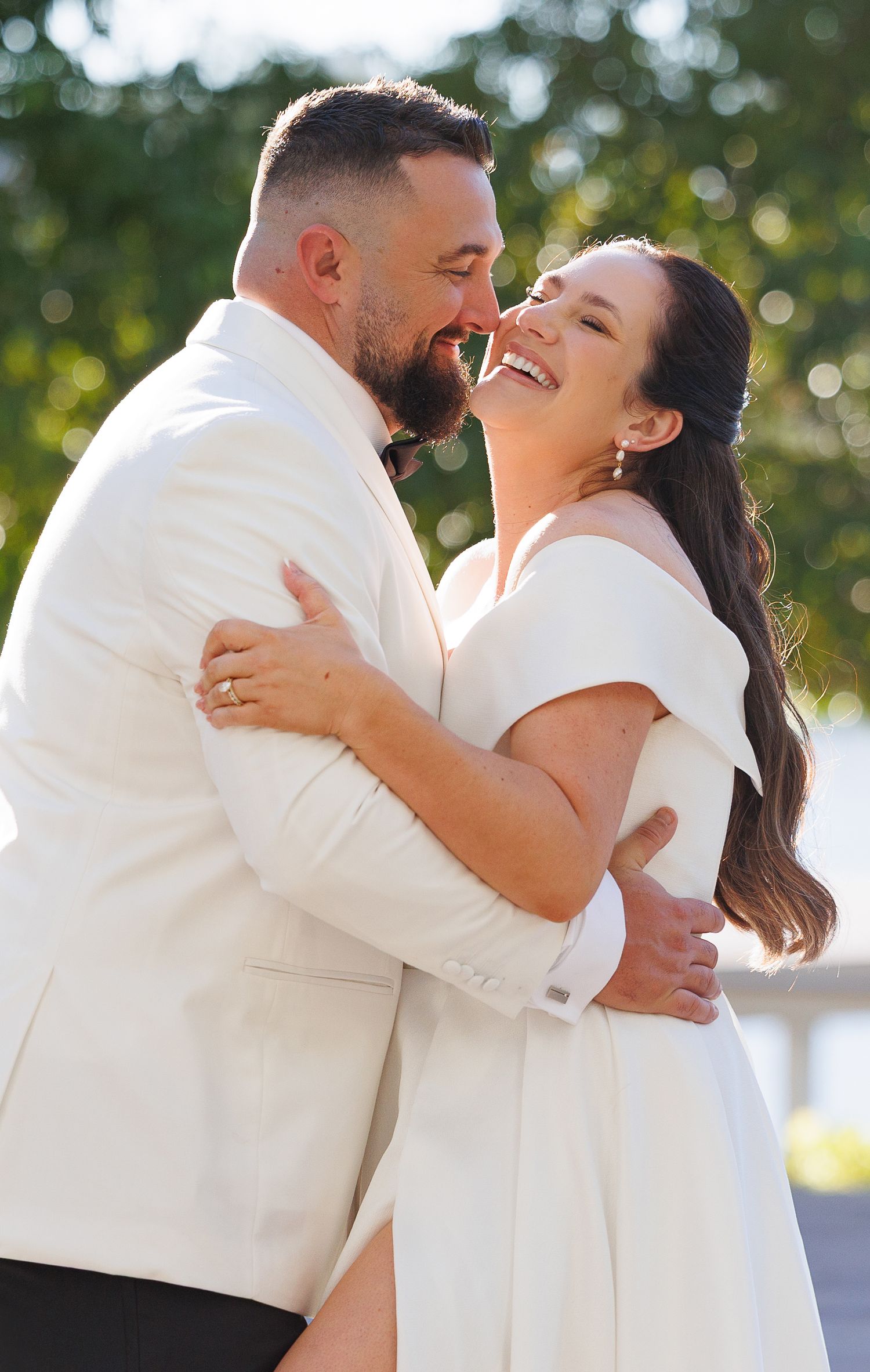 Joyful bride and groom in white attire embracing and smiling outdoors on their wedding day.