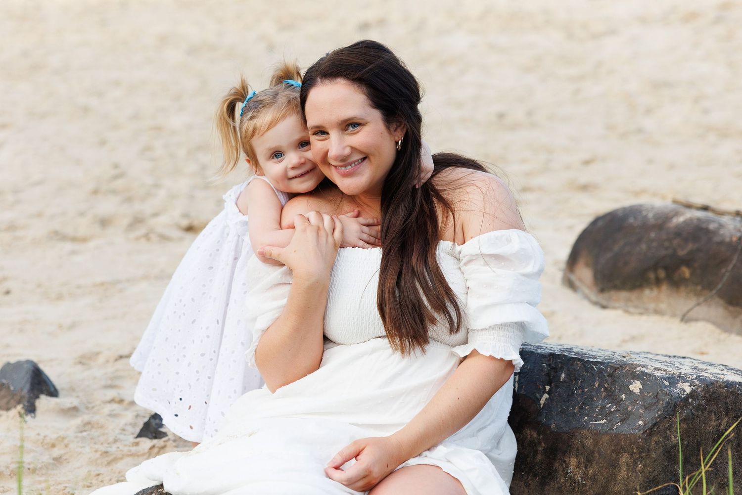 A woman and young girl in white dresses smile together on a sandy beach, sharing a warm embrace.