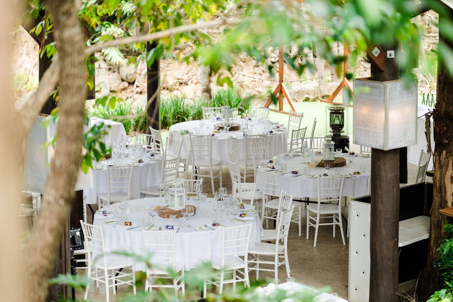 Elegant outdoor wedding reception setup with white chairs and round tables under trees in natural lighting.