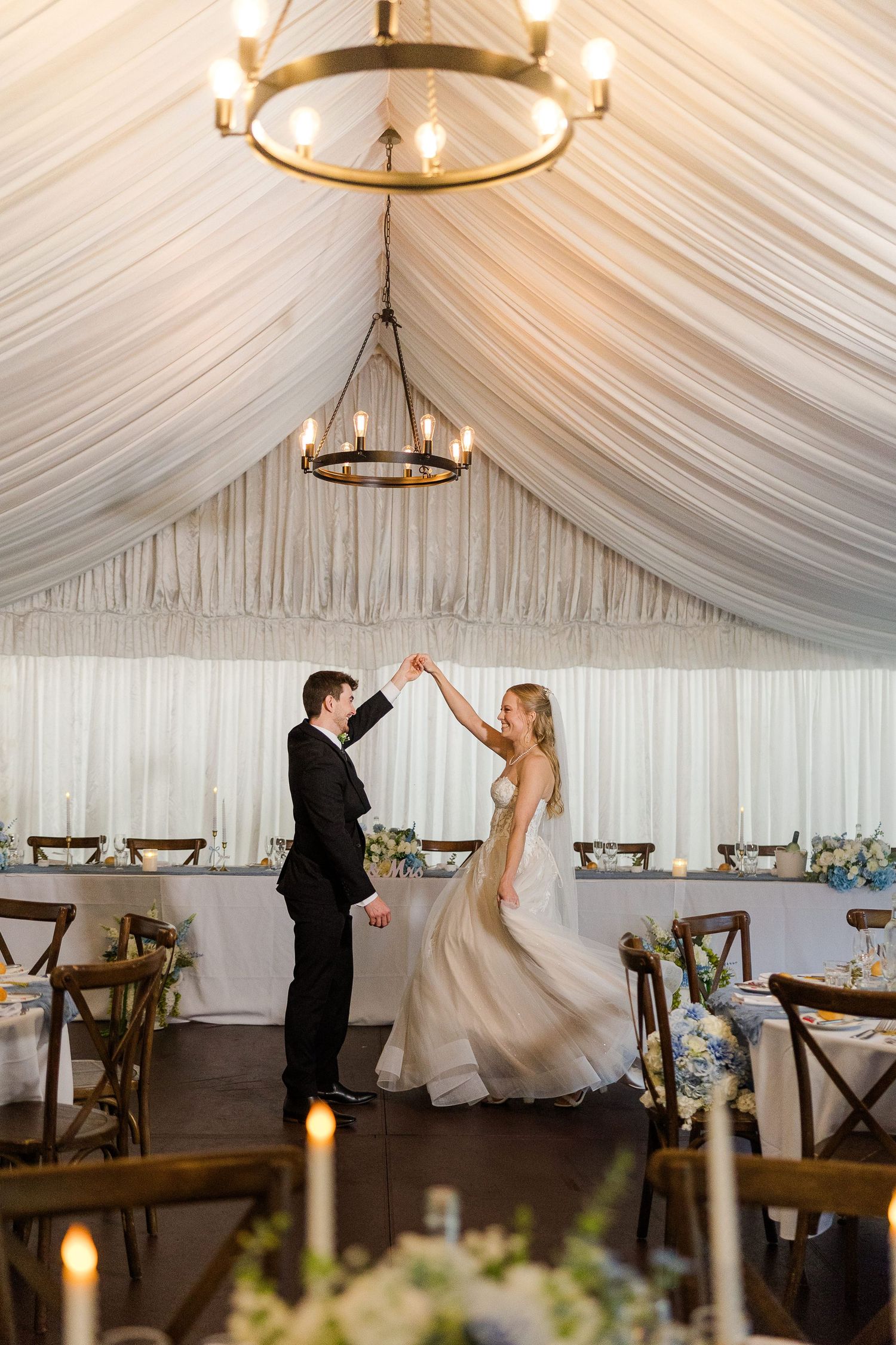 Bride and groom share first dance under elegant white draped tent with chandelier lighting at romantic wedding reception.
