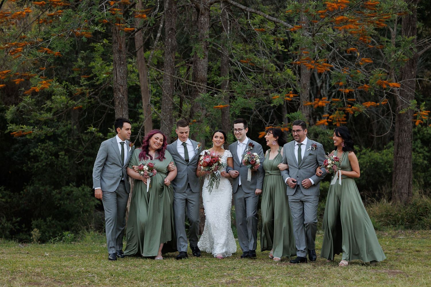 Wedding party walks outdoors in fall forest, bridesmaids in sage green gowns and groomsmen in gray suits flanking bride and groom.