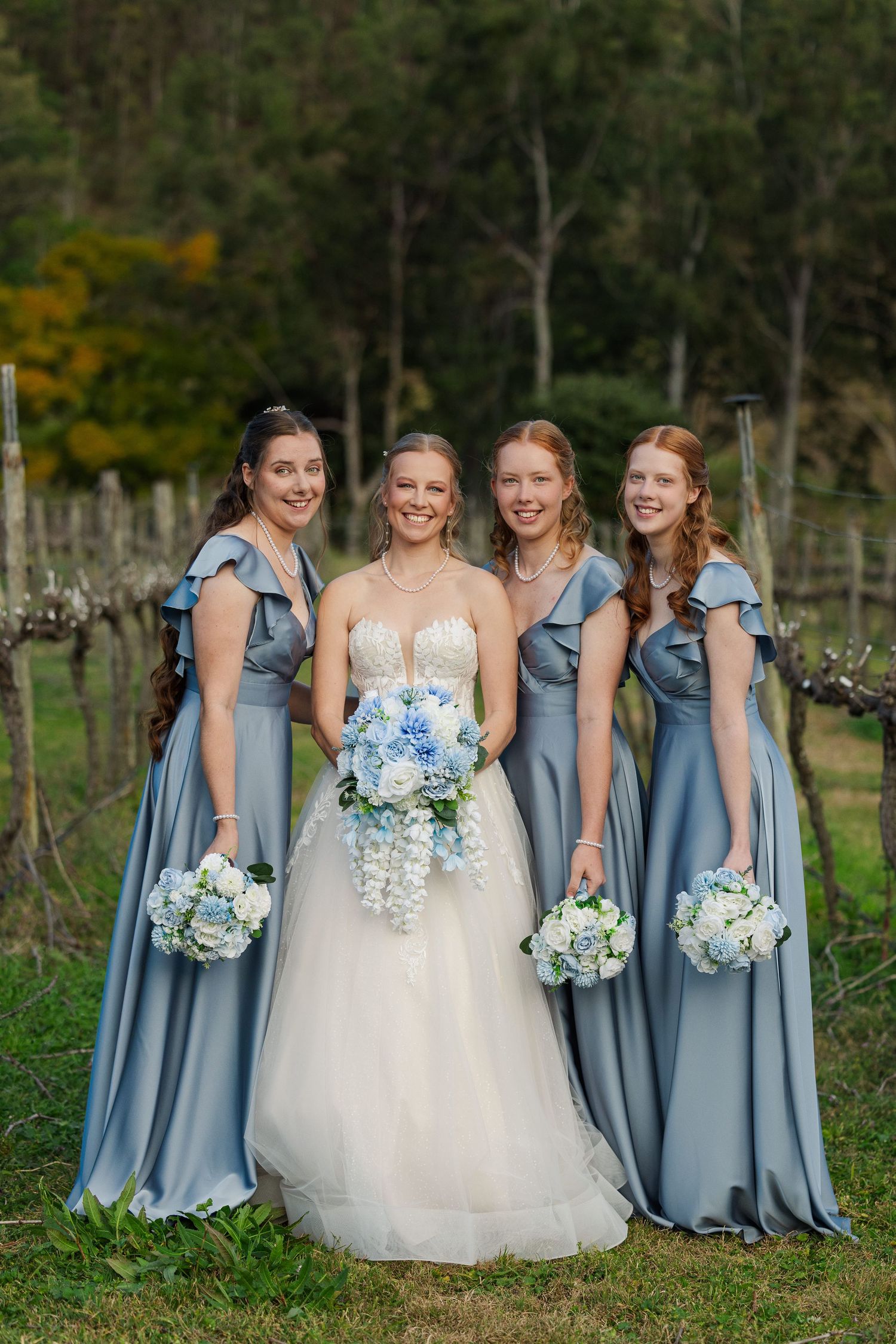 Bride in white gown with three bridesmaids in blue dresses holding floral bouquets at a vineyard wedding.