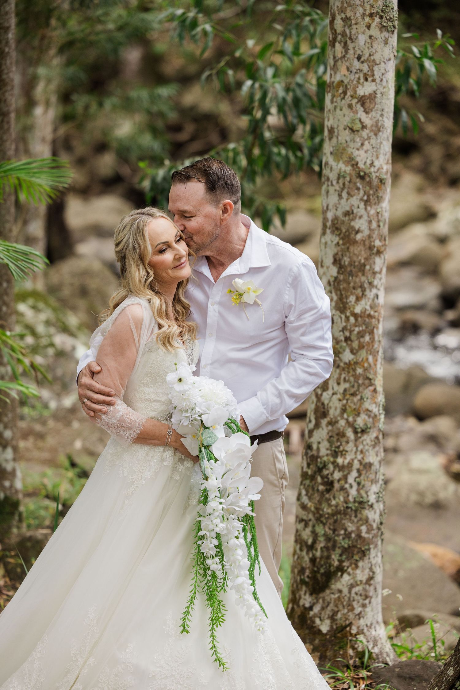 Bride and groom share a romantic kiss among forest trees, bride holding white cascading bouquet in wedding attire.