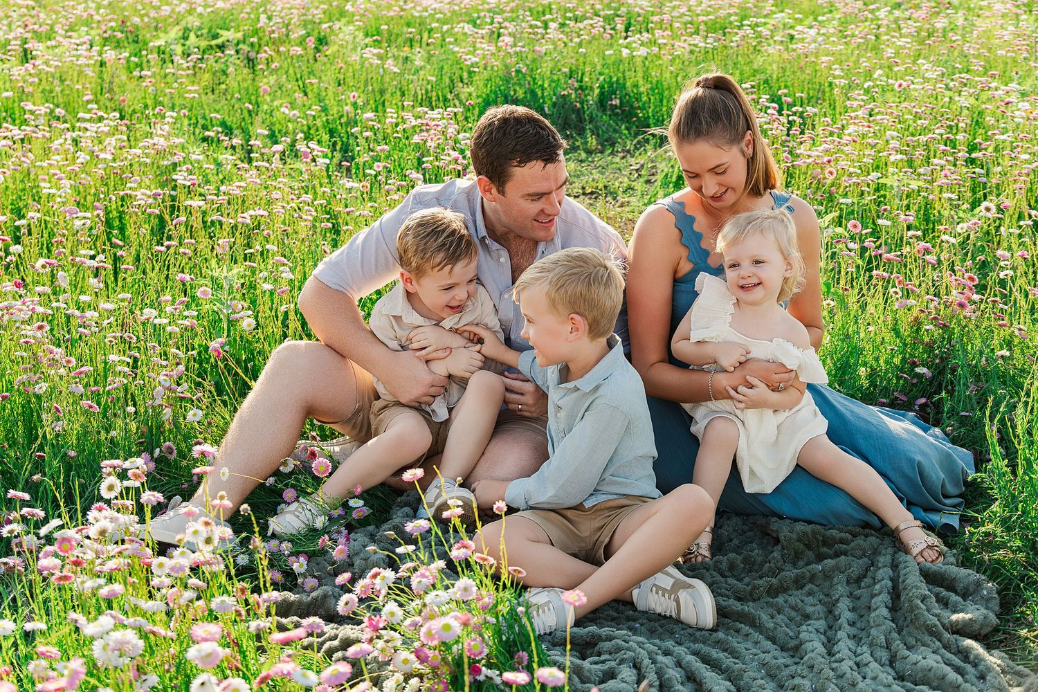Happy family of five sitting together on a blanket in a sunny wildflower meadow, with three young children enjoying a summer day outdoors.