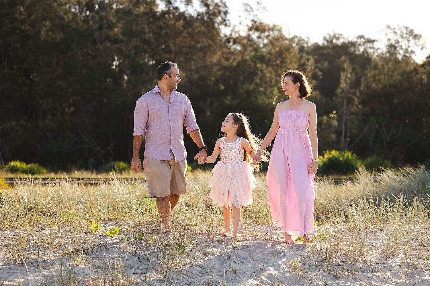 A family walks together holding hands through a sunlit meadow at golden hour, wearing coordinated pink and neutral outfits.