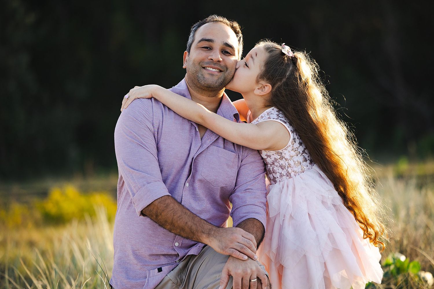 A loving couple embraces in a sunny field during golden hour, with the woman wearing a pink dress and kissing her partner.