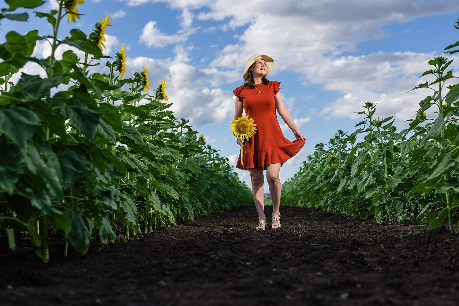 A person in flowing red dress stands in a field pathway between tall green crops under a blue cloudy sky.
