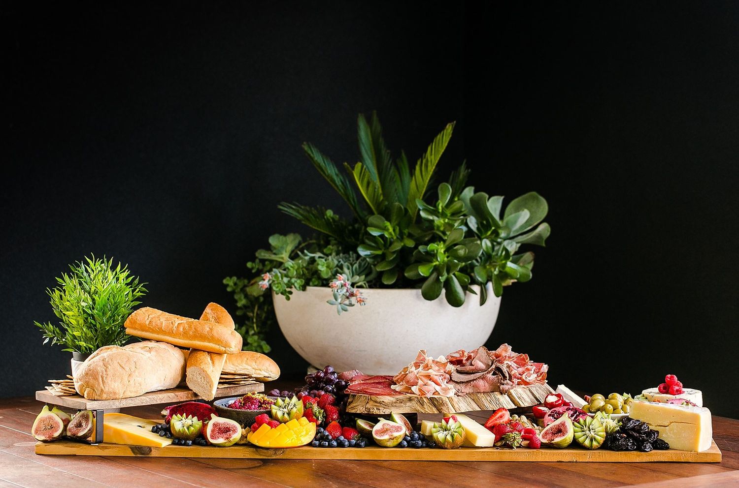 Wooden charcuterie board with meats, cheeses, bread, and fresh herbs against dark background with potted plants.