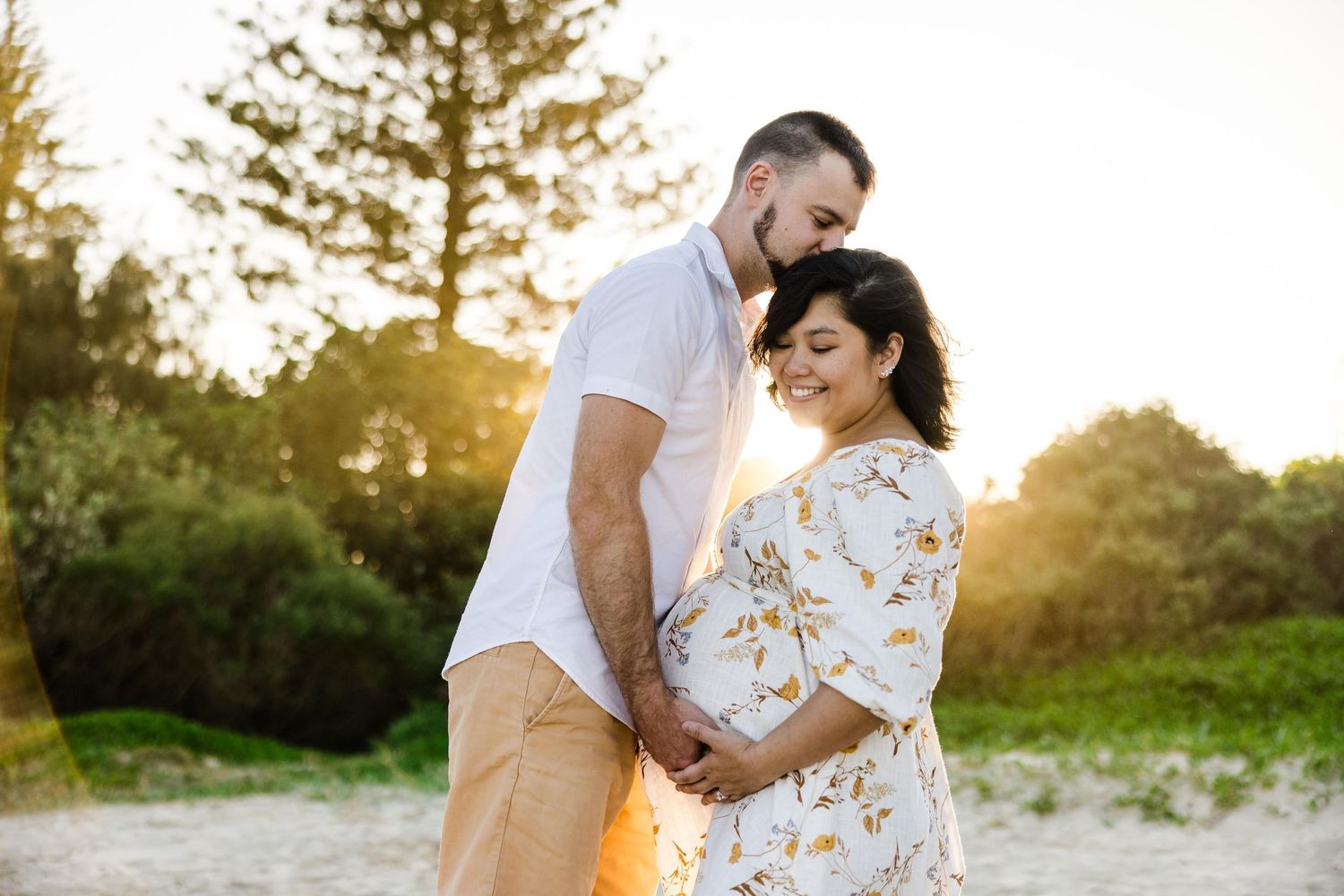 A couple shares an intimate moment during sunset on a beach, with golden light filtering through trees in the background.