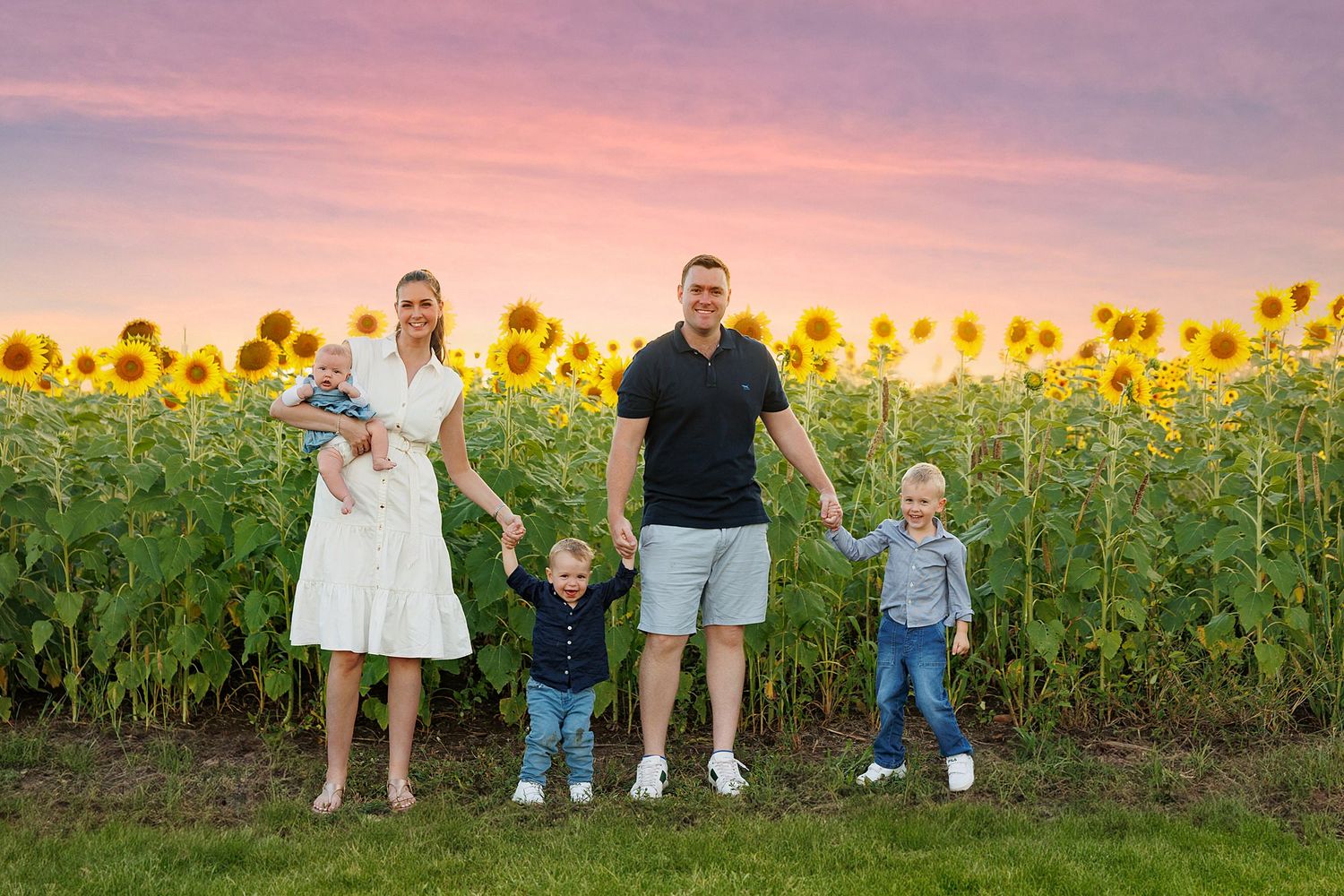 Group stands together by a vibrant sunflower field at sunset with pink and purple sky.