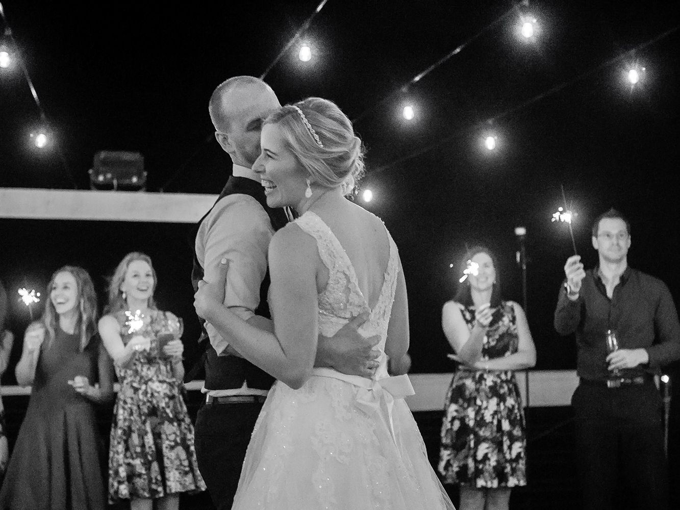 Black and white photo of newlyweds dancing at reception with string lights overhead.