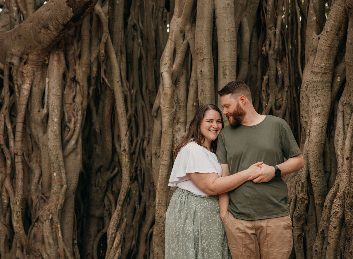 A couple stands together in front of large tree roots during a romantic outdoor photoshoot.