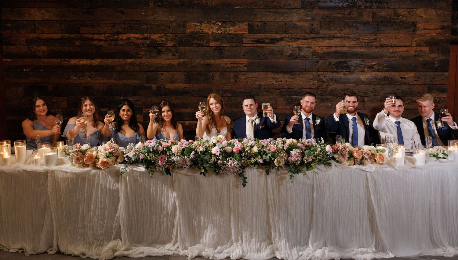 A wedding party sits at an elegant head table with white draping and floral centerpieces against a rustic brick wall.