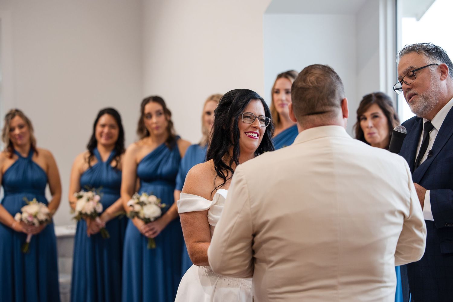 Bride smiling at groom during wedding ceremony with bridesmaids in blue dresses holding white bouquets in background.