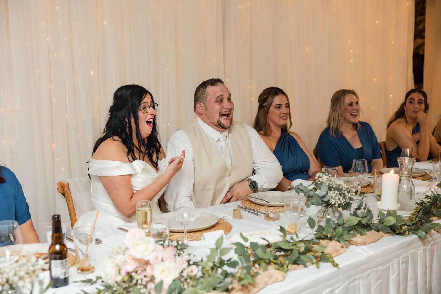 Bride and groom laughing joyfully at wedding reception head table with bridesmaids in blue dresses and floral arrangements.