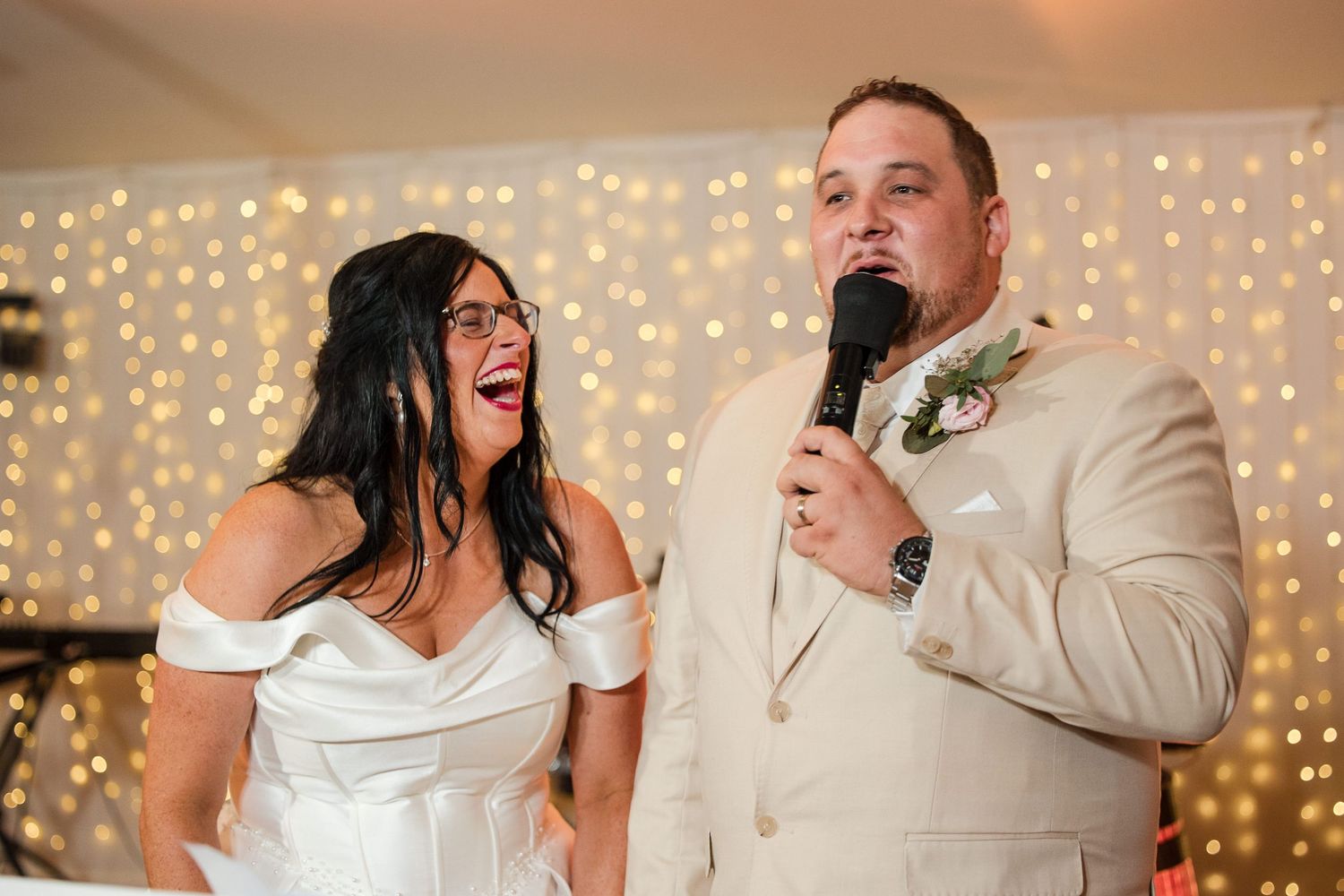 Bride and groom laughing and speaking into microphone at wedding reception with twinkling lights backdrop.