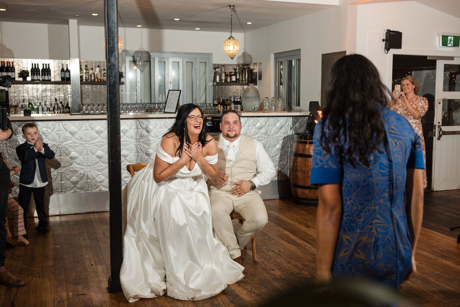 Bride and groom seated together laughing during wedding reception at elegant bar venue with guests watching.