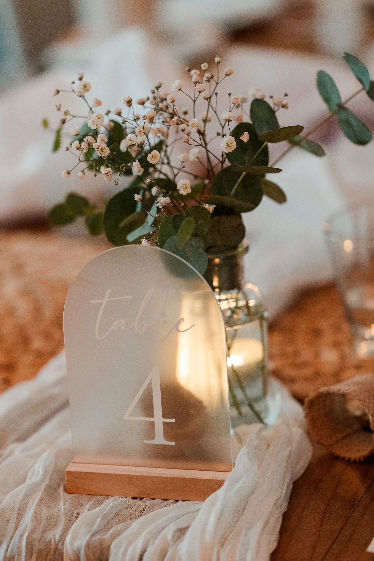 Frosted acrylic arch table number 4 sign with LED light beside eucalyptus and baby's breath floral arrangement.