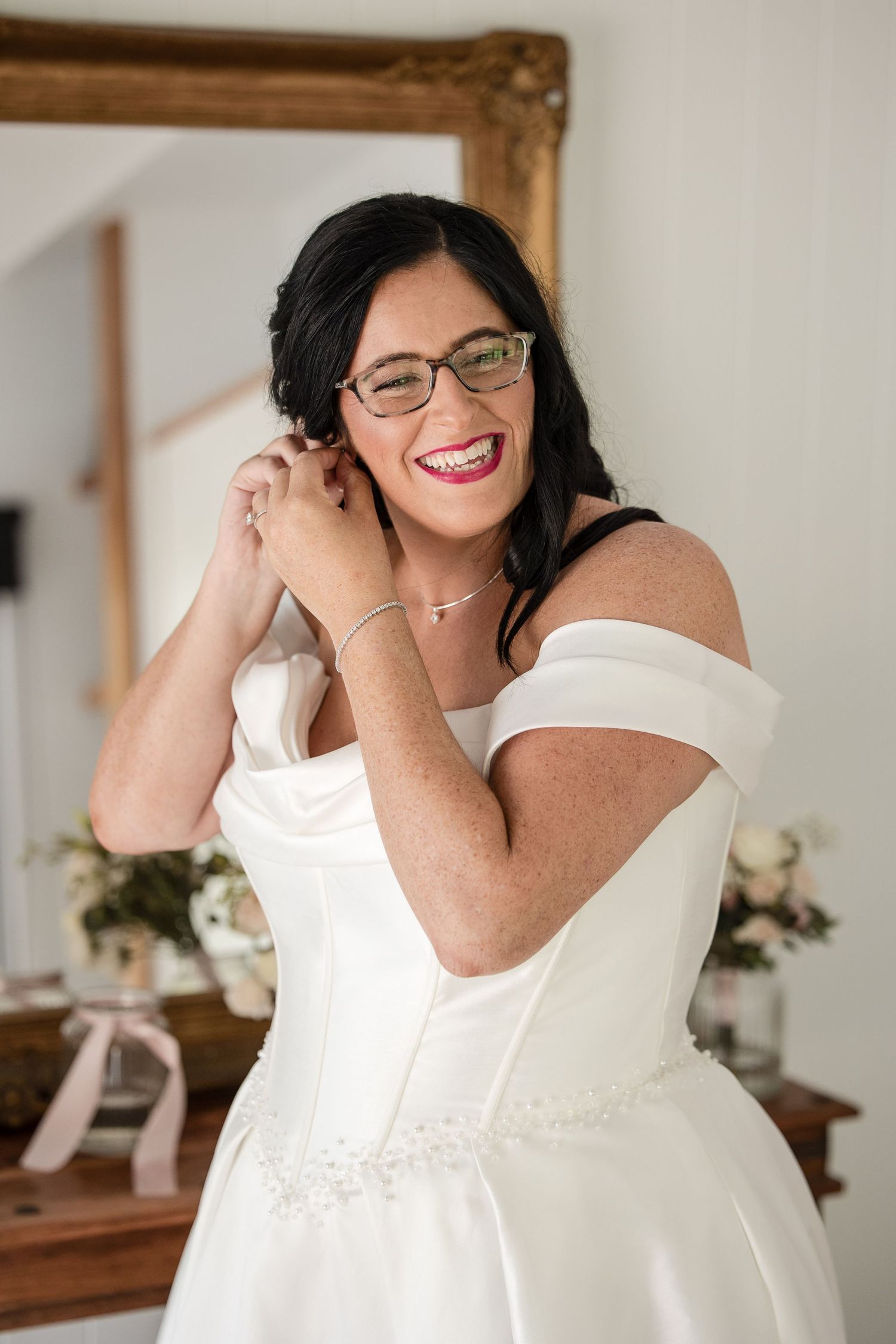 Smiling bride in white off-shoulder gown and glasses adjusting earring before gold mirror.
