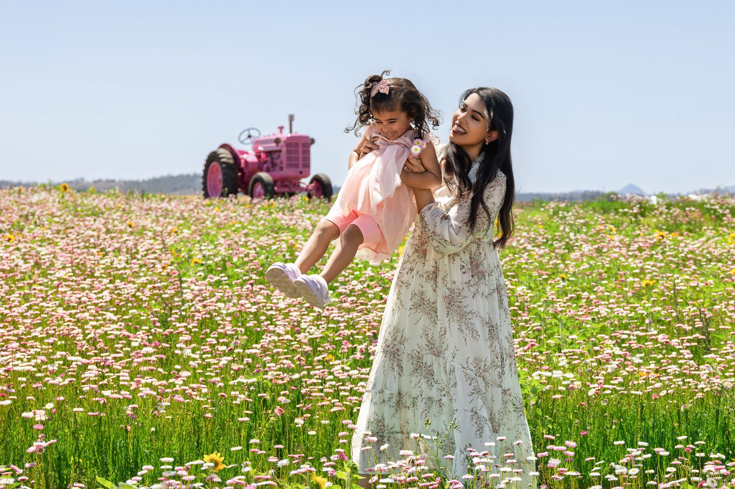 Woman in floral dress lifting toddler in pink outfit in a daisy field with a pink tractor in the background.
