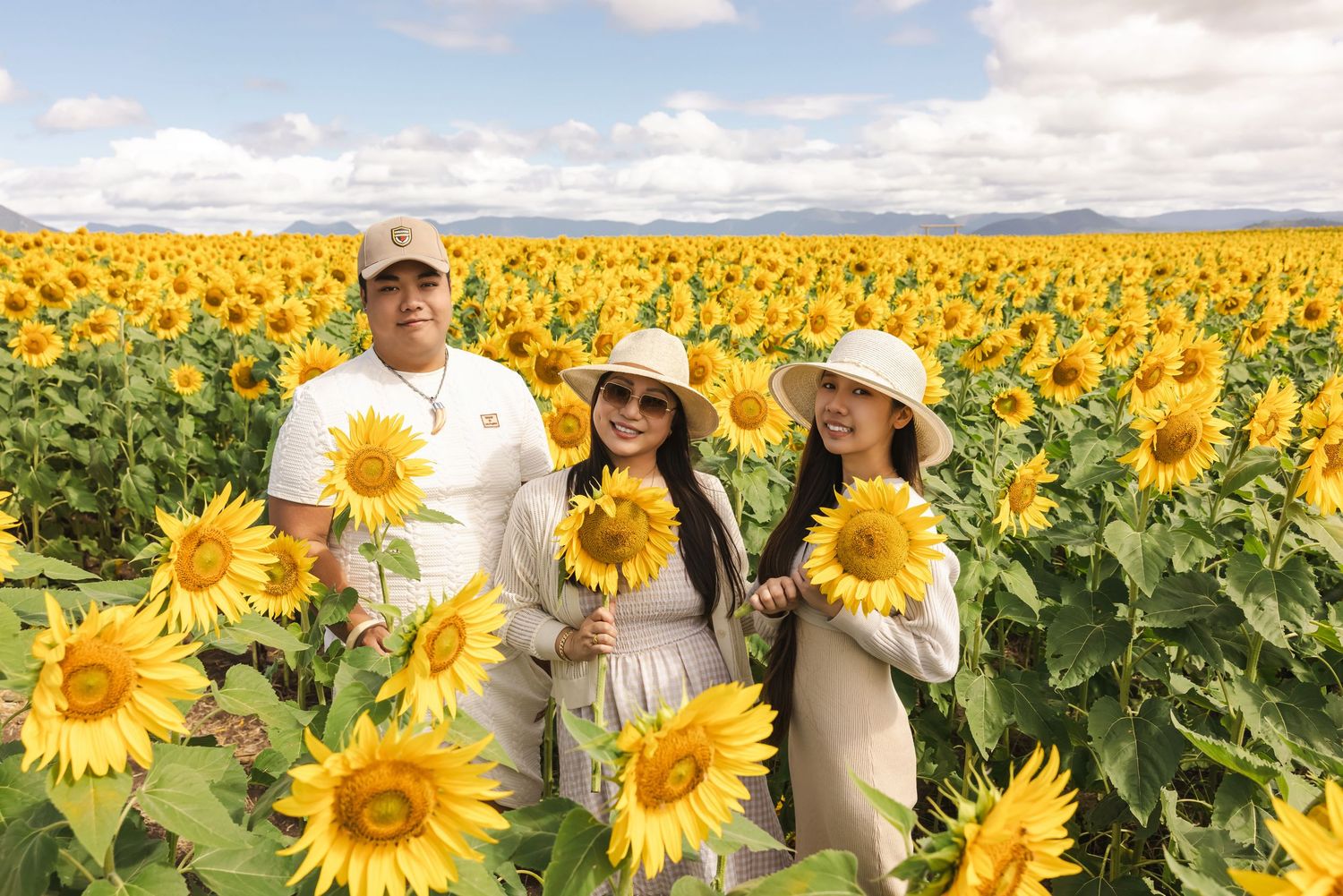 Three people posing happily in a vast sunflower field under a partly cloudy sky, holding bright yellow sunflowers.