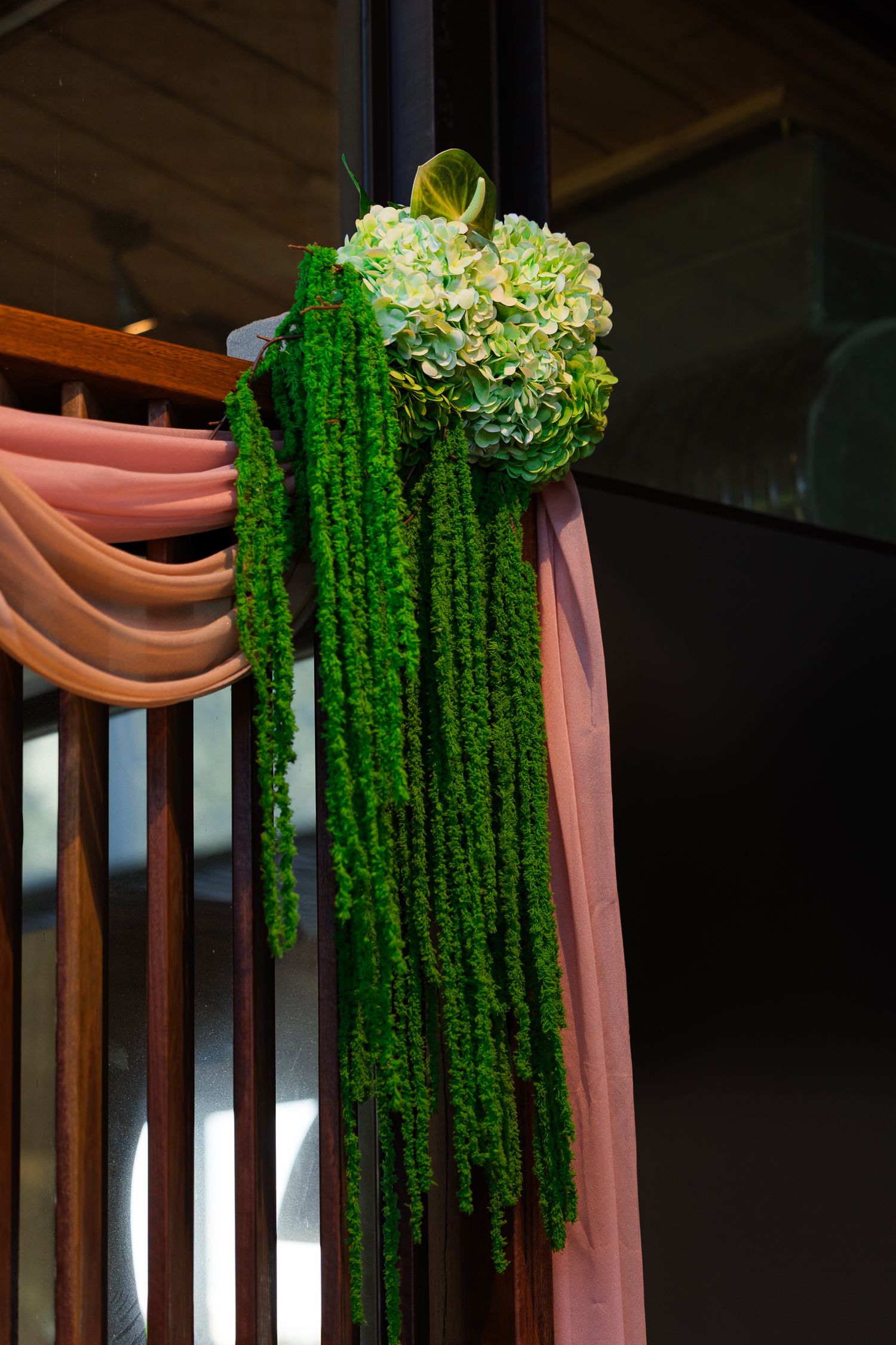 Green hydrangea and cascading amaranthus floral arrangement with pink fabric draped on wooden railing.