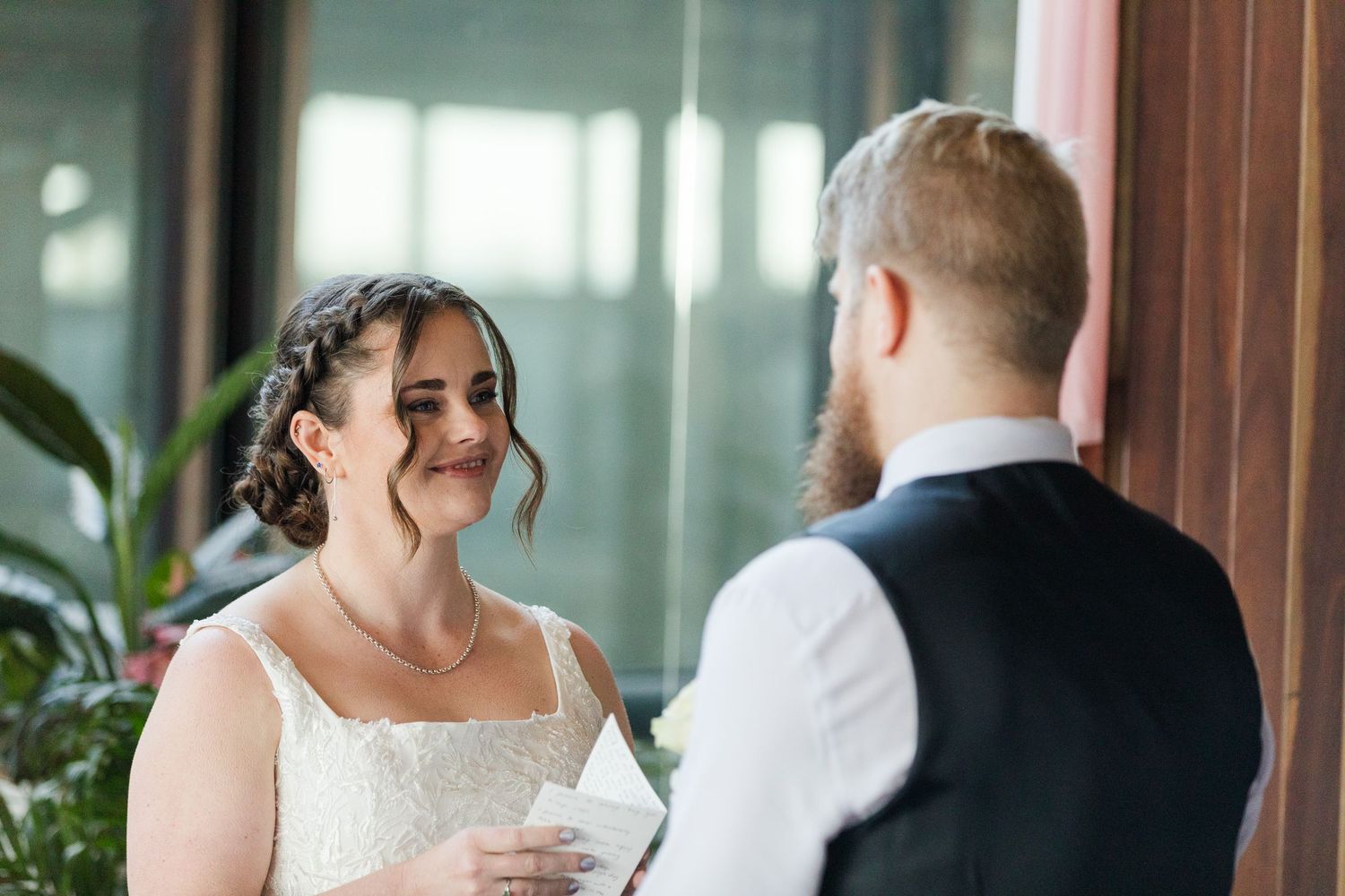 Bride smiling at groom during wedding ceremony vows indoors with greenery background.