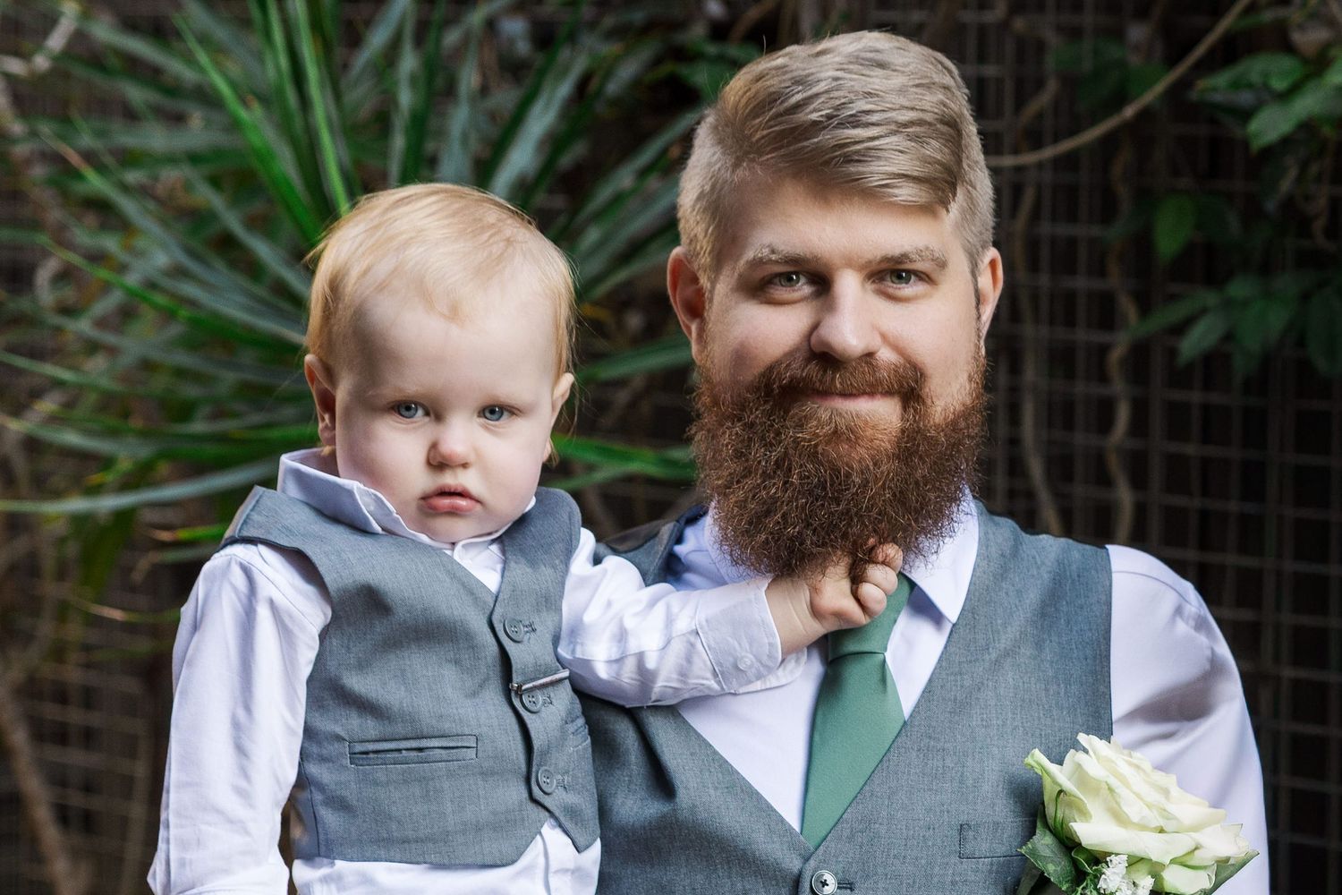 Bearded man in gray vest and green tie holds a young toddler boy in matching vest at a wedding.