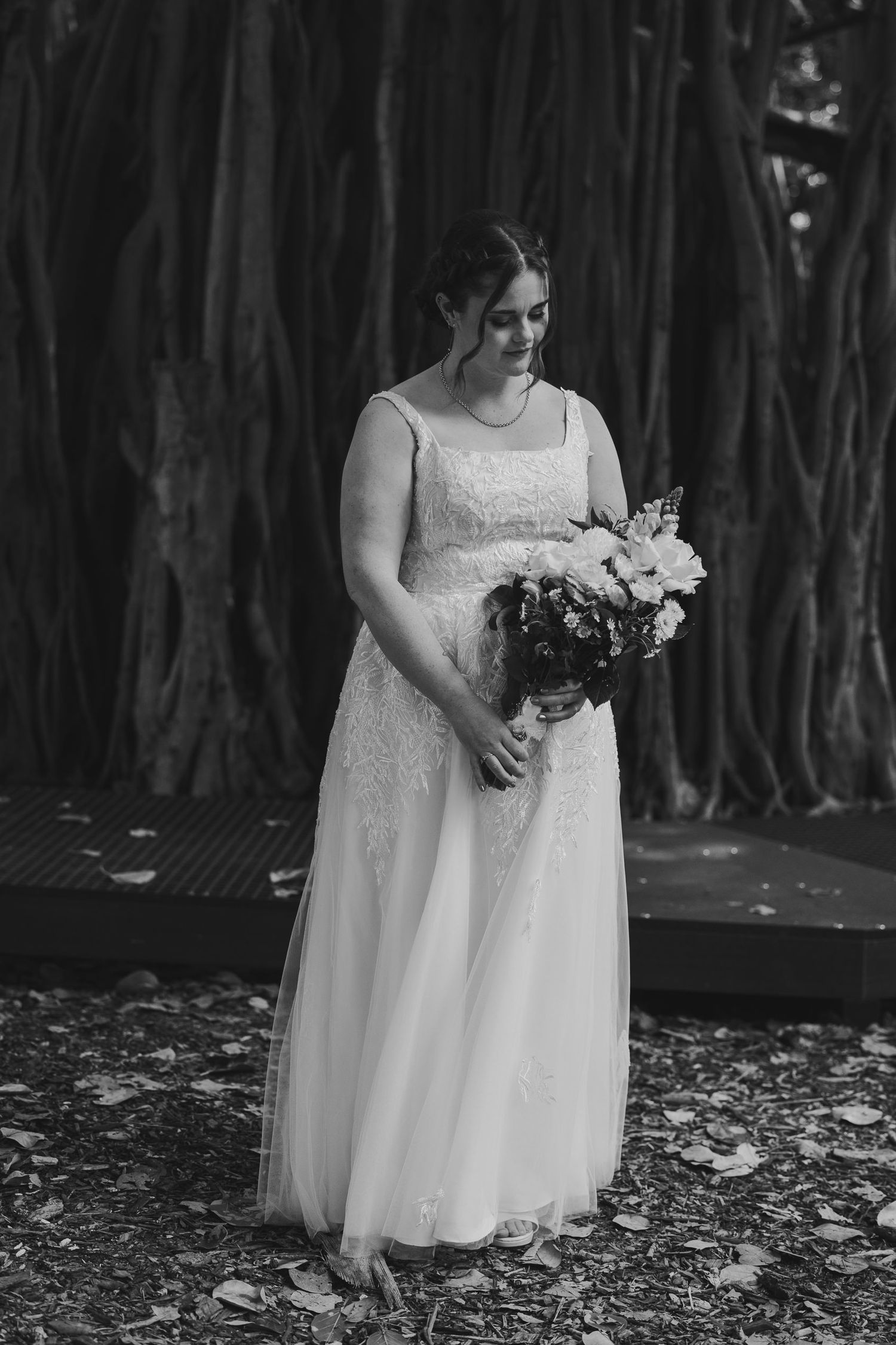 Bride in white lace gown holding floral bouquet standing before banyan trees in black and white wedding photo.