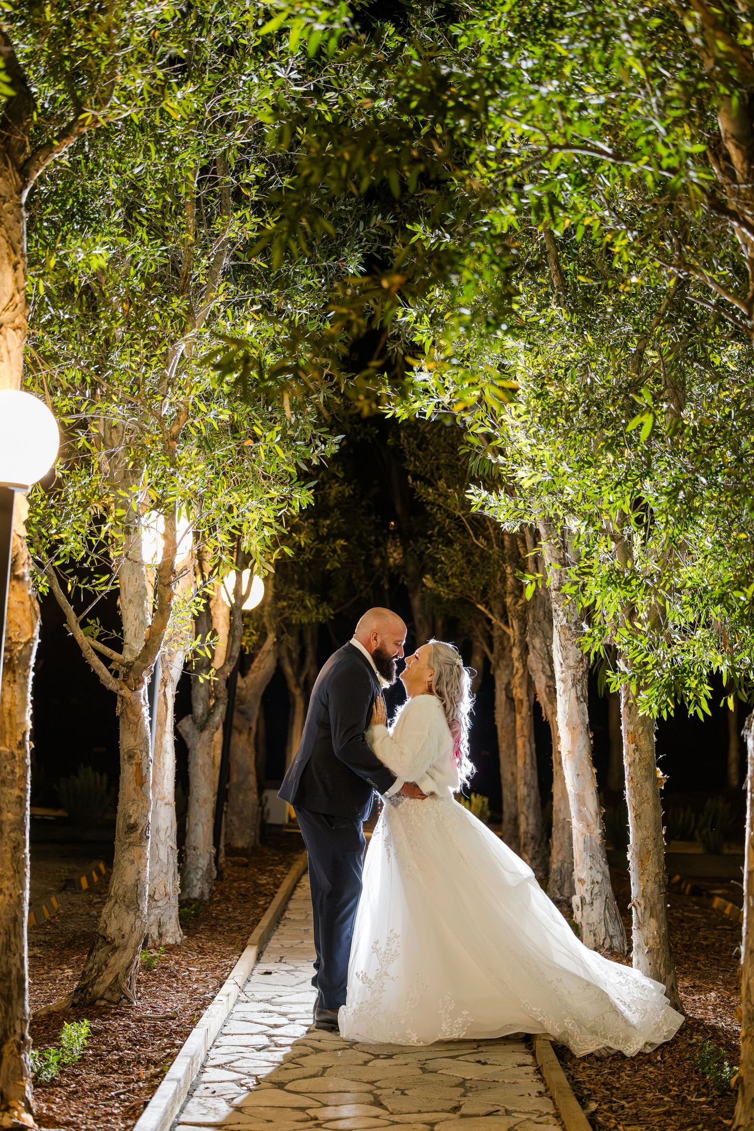 Bride and groom share a romantic kiss under a canopy of illuminated trees on a stone pathway at night.