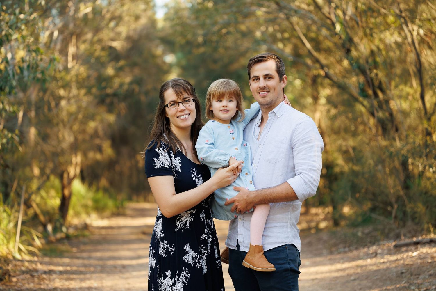 A family stands together on a sunlit tree-lined path during an autumn outdoor photo session.
