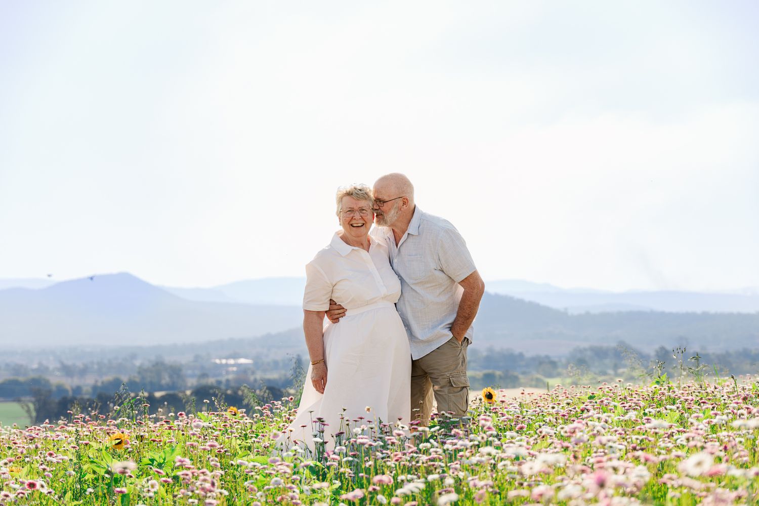 Elderly couple embracing in a wildflower meadow with scenic mountain views on a sunny day.