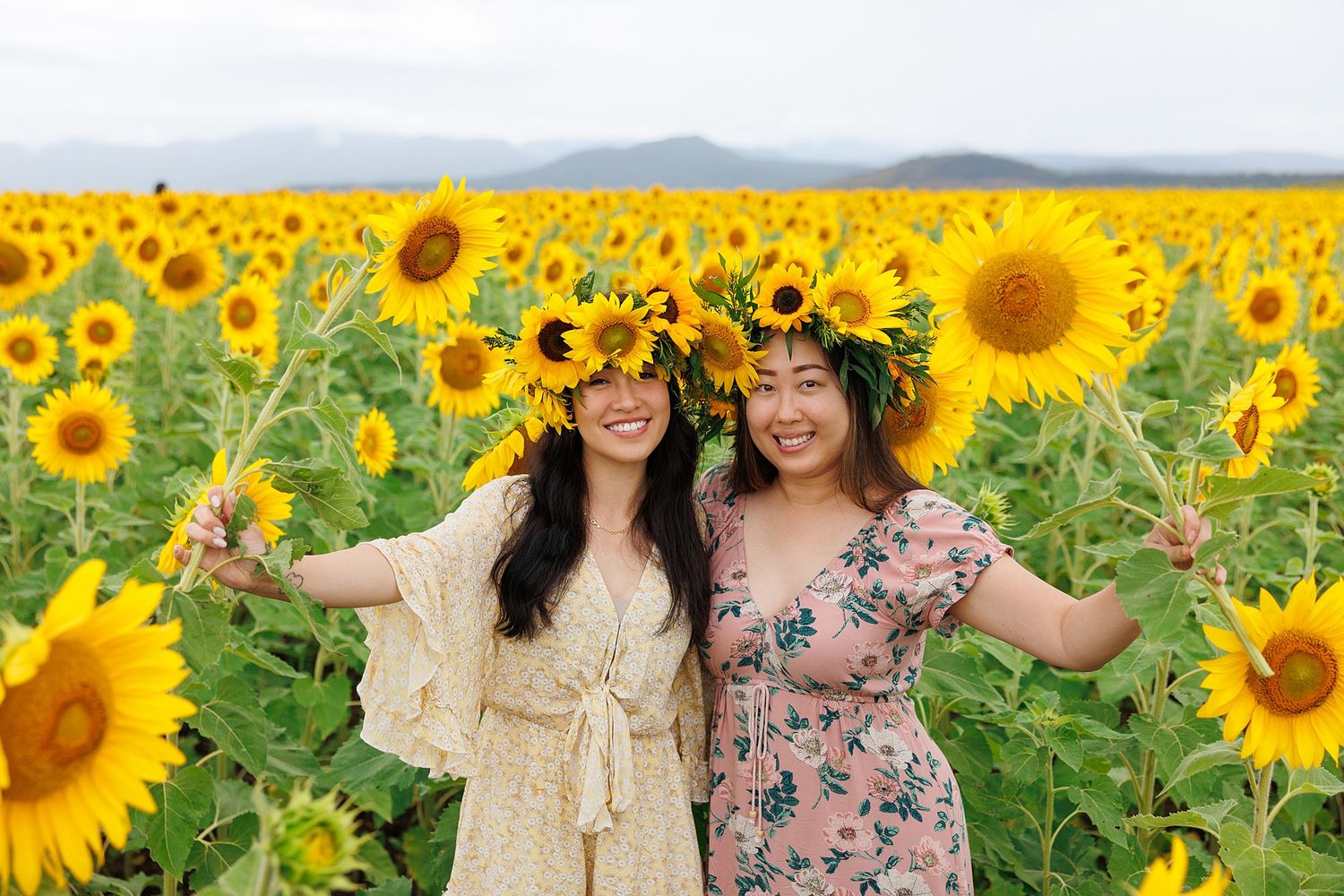 Two people wearing floral crowns smile together in a vibrant field of blooming yellow sunflowers on a sunny day.