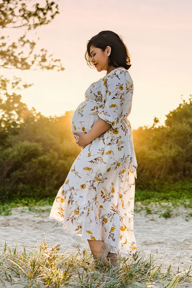 A woman in a floral dress stands in golden sunlight during a maternity photoshoot on sandy ground.