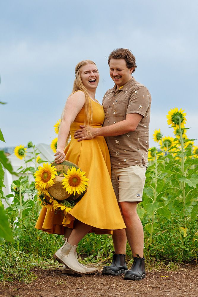 Happy couple laughing in sunflower field with woman in yellow dress holding flowers.