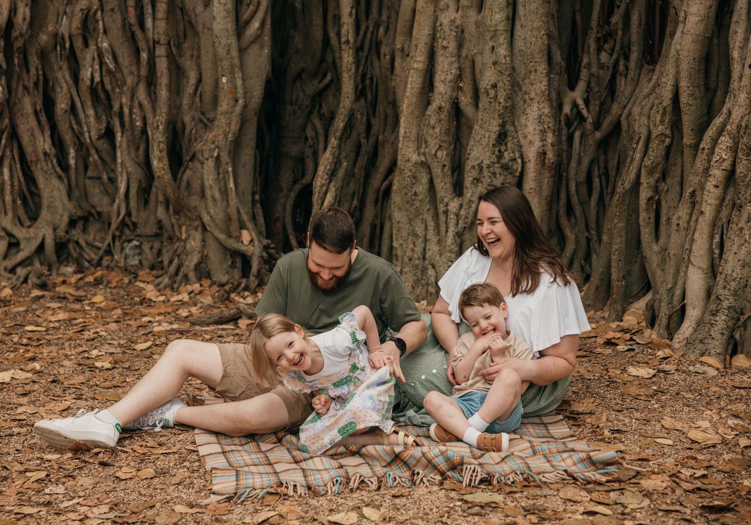 A family sits together on fallen leaves under a large banyan tree with dramatic aerial roots in a natural setting.