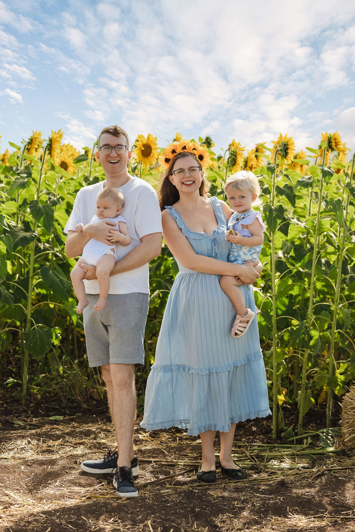 Smiling family with two babies posing in front of a blooming sunflower field under blue sky.