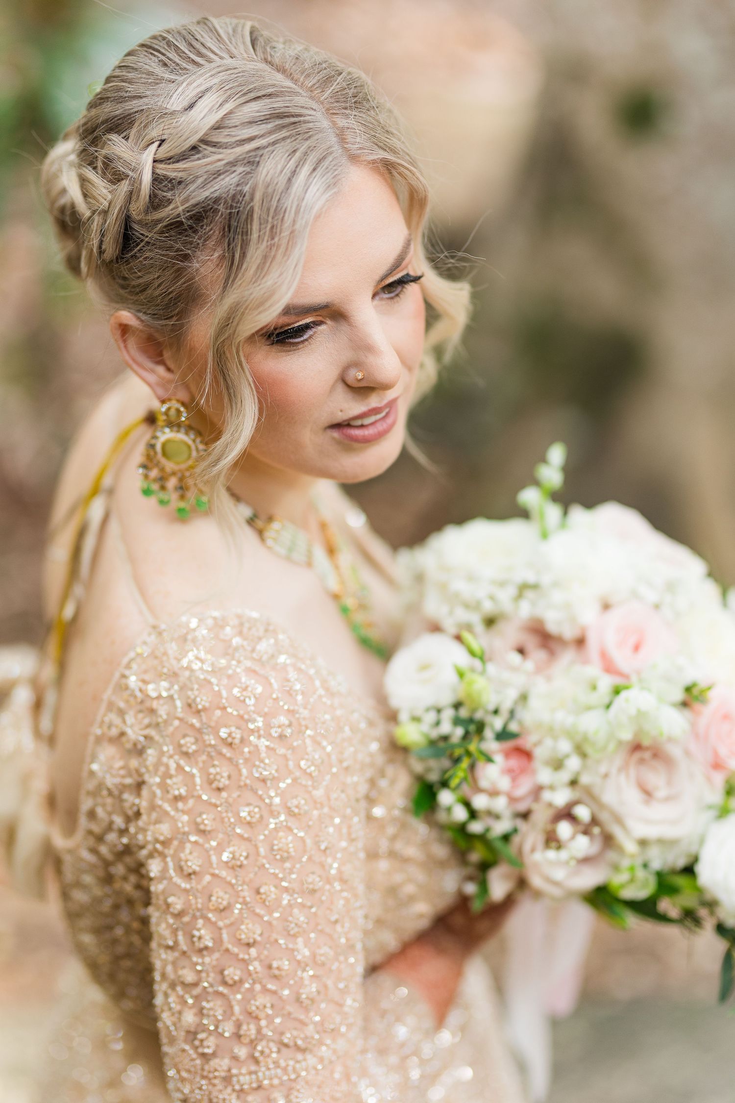 Elegant bride in gold beaded gown holding blush and white floral bouquet with braided updo hairstyle.