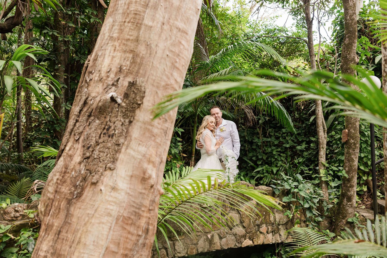 Couple embracing on a stone bridge surrounded by lush tropical greenery and ferns in a garden setting.