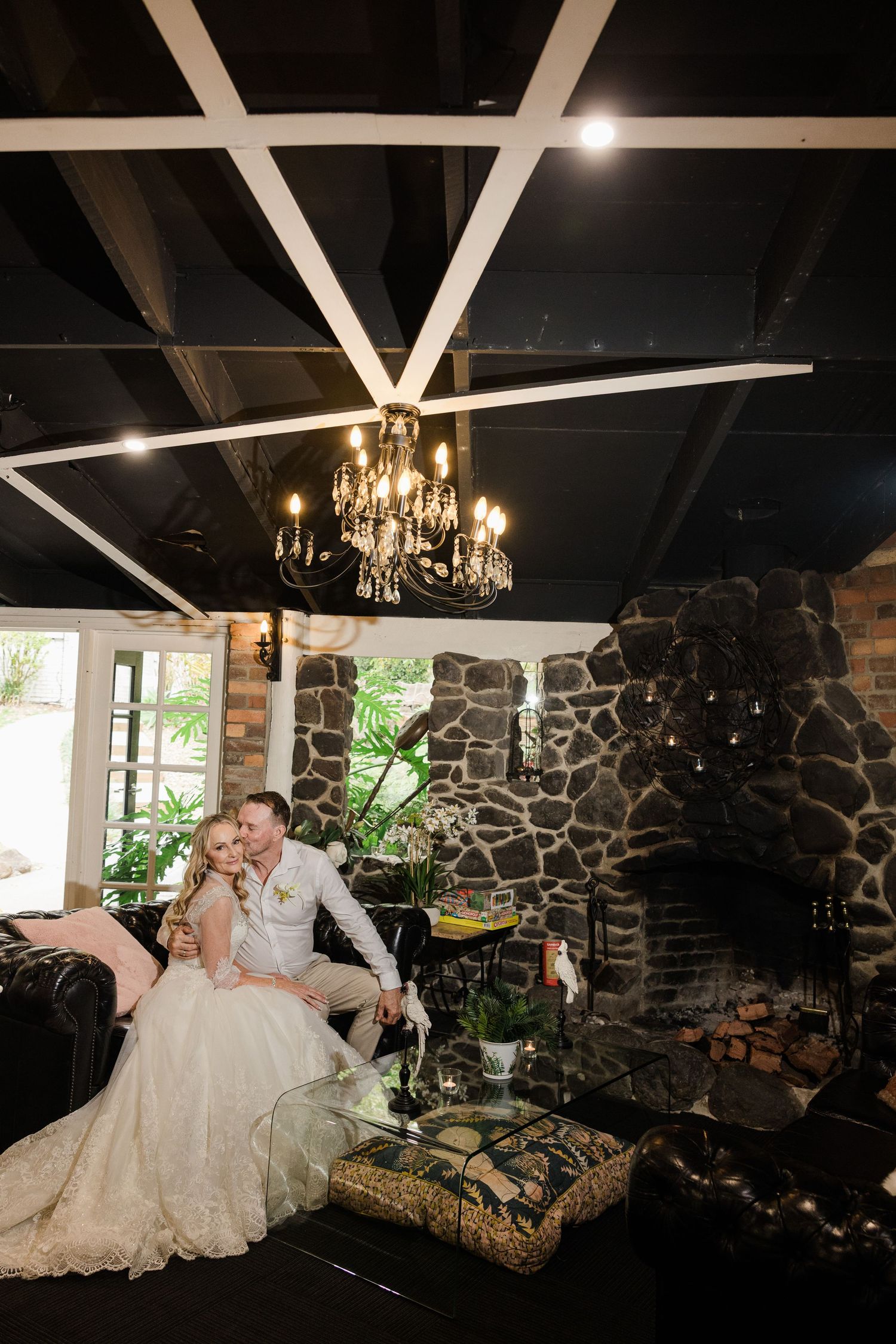 Bride and groom sitting together by a stone fireplace under a chandelier in an elegant wedding venue.