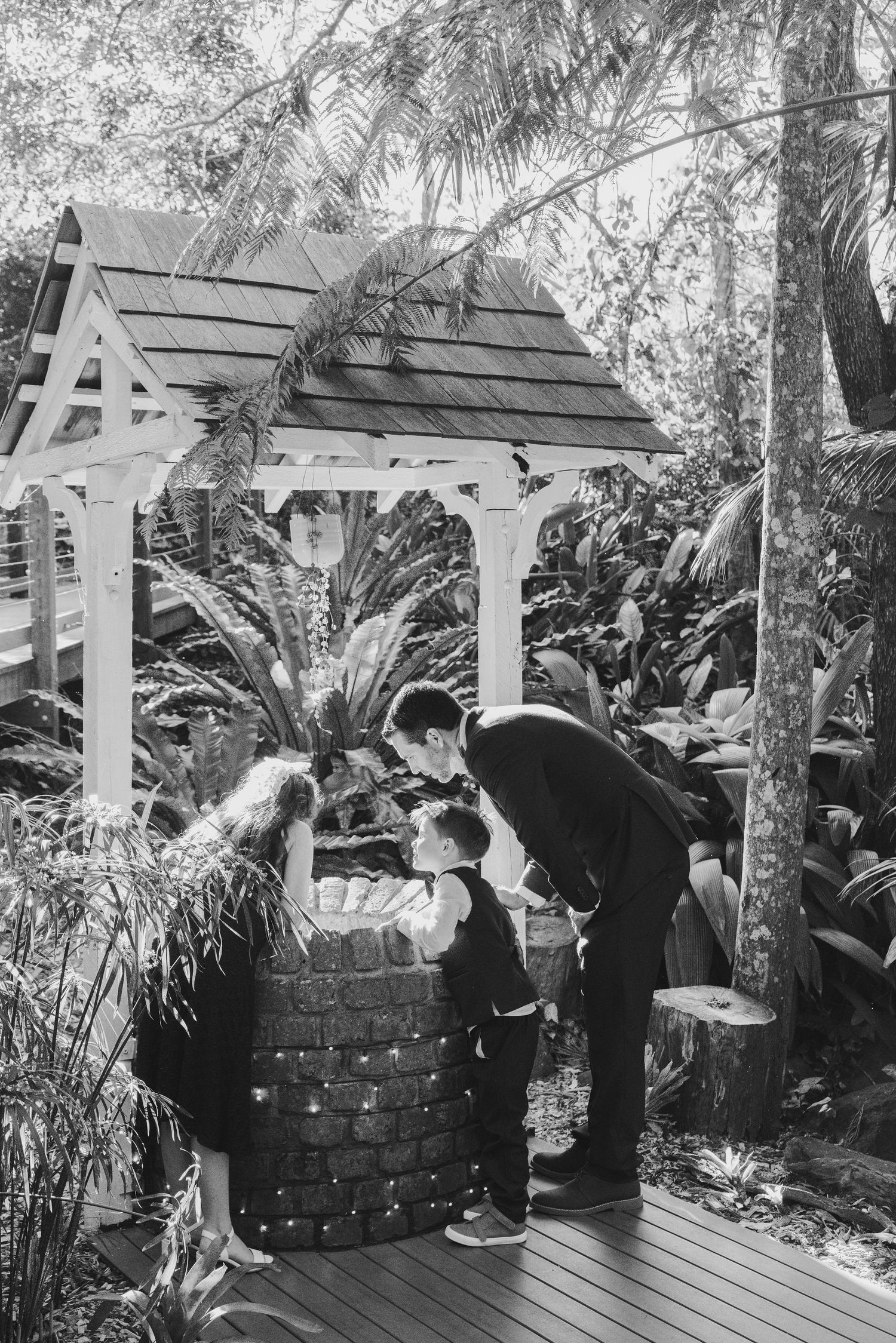 A couple stands beneath a wooden garden arbor surrounded by lush tropical plants in black and white.