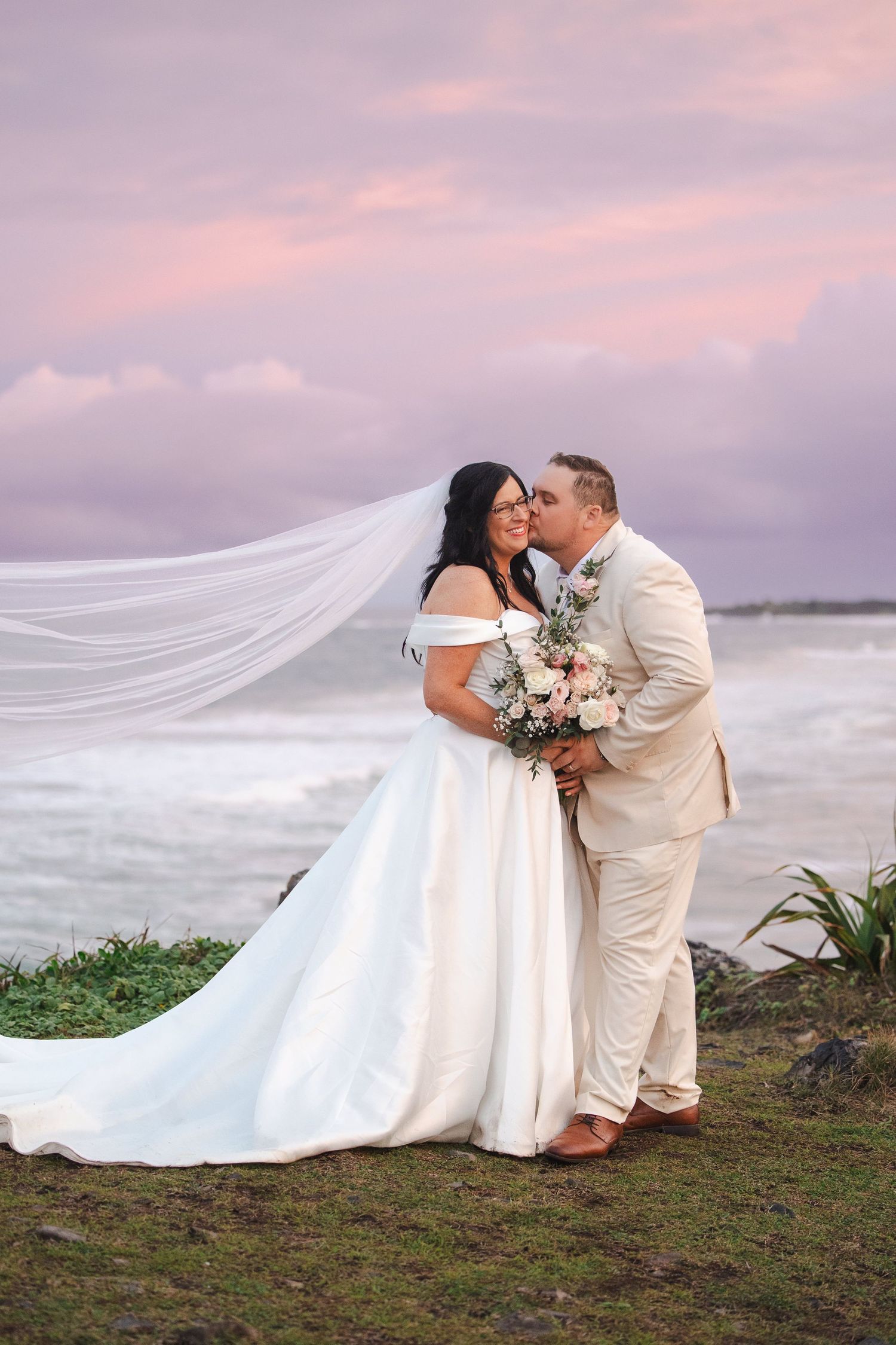 Bride and groom share a kiss at sunset on a coastal cliff, her veil flowing in the wind, wearing white gown and tan suit.