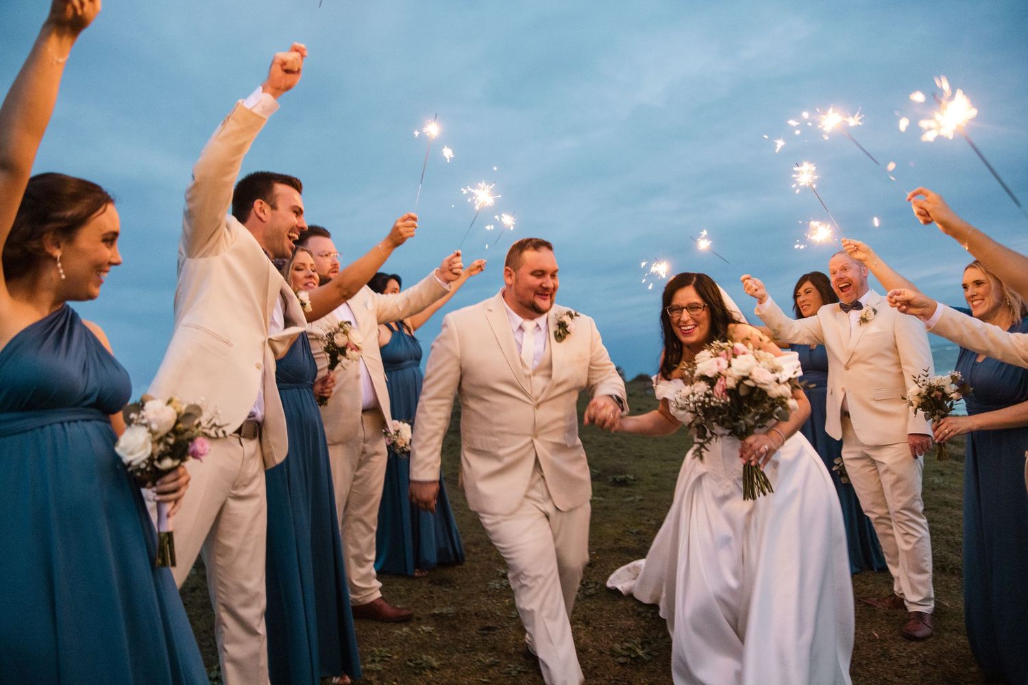 Bride and groom walk through sparkler tunnel with wedding party in blue dresses and tan suits at outdoor evening ceremony.