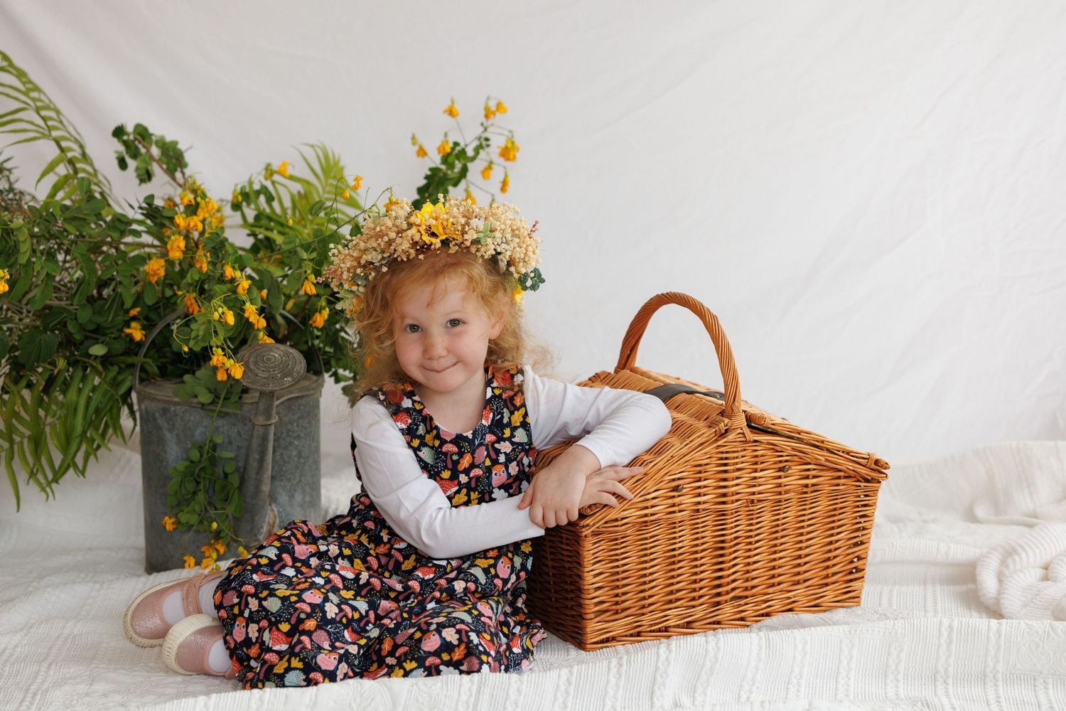 Smiling curly-haired girl in floral dress and flower crown sits beside wicker basket and yellow wildflowers.