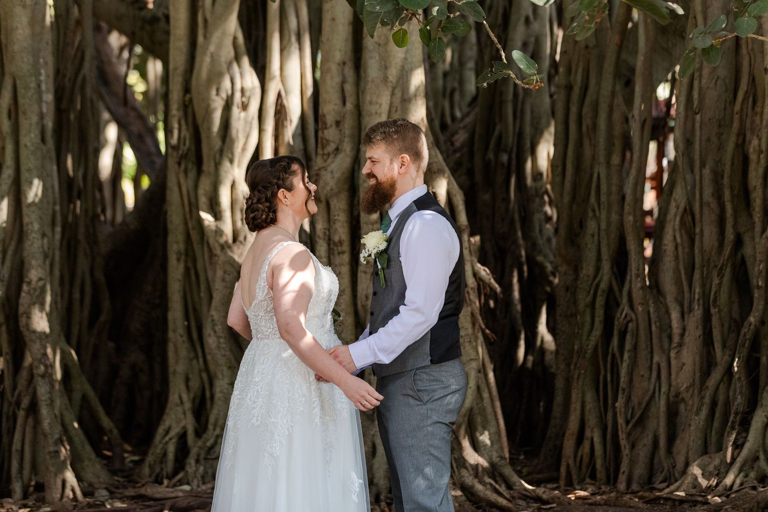 Bride and groom holding hands, smiling at each other in front of large banyan trees on their wedding day.