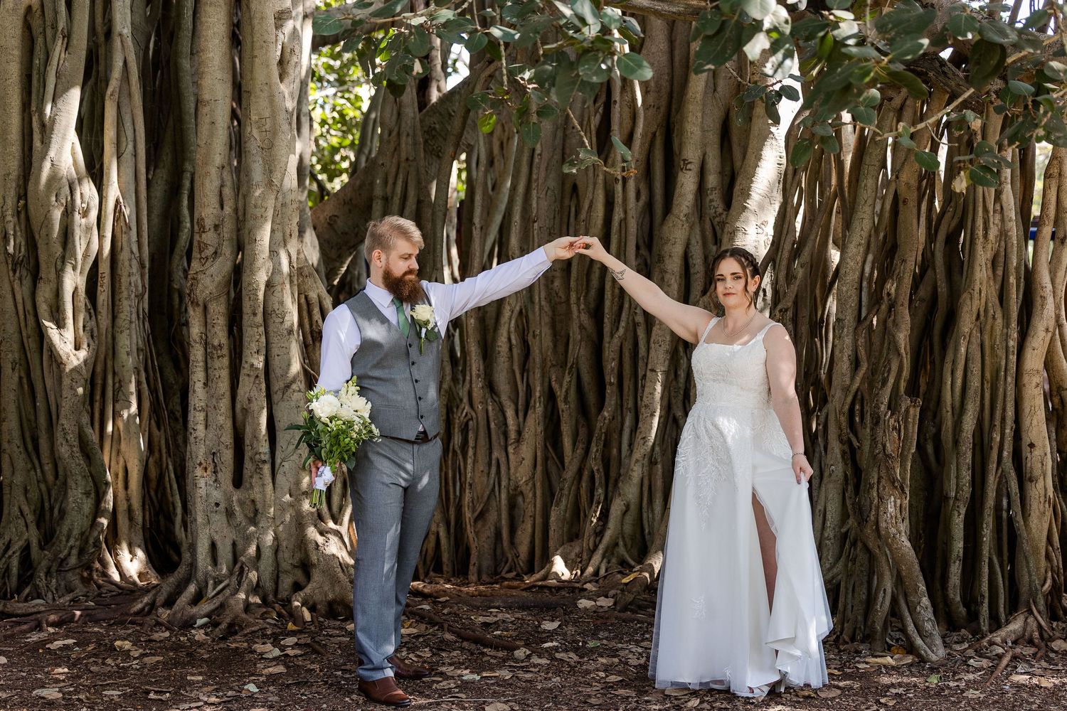 Bride and groom hold hands under a grand banyan tree, bride in white gown, groom in grey vest, outdoor wedding.